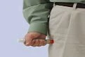 A hand holding a syringe horizontally near the back pocket of a pair of light-colored pants worn by a person with a green shirt