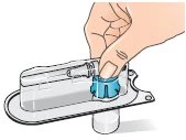 One hand presses the blue button on a transparent medical device placed on a flat base to activate the internal mechanism