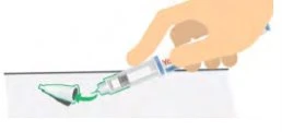 A hand holds a syringe tilted to draw green liquid from a glass vial on a white surface
