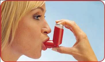Profile of a blonde woman using a red spray inhaler held with the right hand near her open mouth against a light blue background