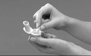 Two hands hold a white medical device while one hand inserts a small circular component into the top part