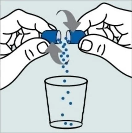 Two hands open a blue capsule, causing it to drop over a transparent glass with small blue granules falling into it
