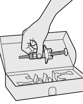 A hand holds a horizontal syringe above an open cardboard box with empty internal compartments for drug containment