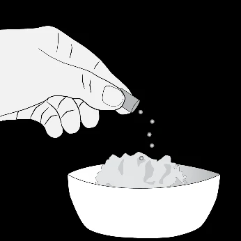 One hand holding a small capsule above a white bowl from which light-colored granules or powder are falling