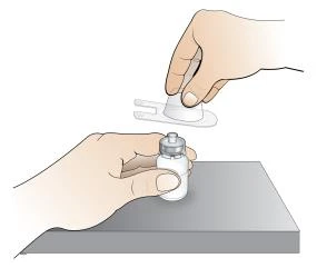 A hand holds a glass vial on a flat surface while another hand prepares a syringe
