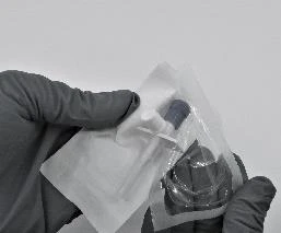 Una mano guantata tiene una fiala medica sigillata all'interno di una bustina di plastica trasparente e bianca su sfondo neutro