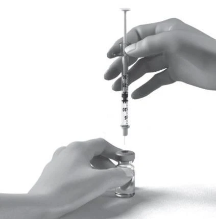 A hand holds vertically a syringe with needle pointing toward a small glass vial held by the