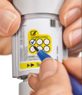 Two hands holding a white medical device while a hand with a blue pen circles an item on a yellow table containing icons and boxes
