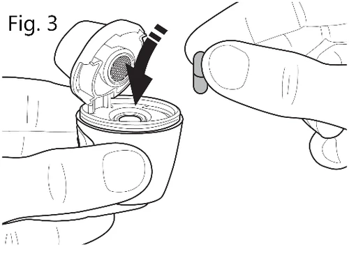 Disegno tecnico che mostra due mani che inseriscono una piccola capsula cilindrica all'interno di un dispositivo medico circolare con una freccia nera