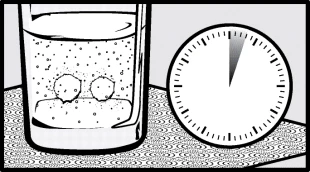 Un bicchiere d'acqua con due compresse effervescenti sul fondo accanto a un orologio analogico che indica l'ora su una superficie a motivi circolari