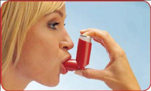 Blondynka trzymająca czerwony inhalator przy ustach i biorąca z niego lek na tle jasnego błękitnego nieba