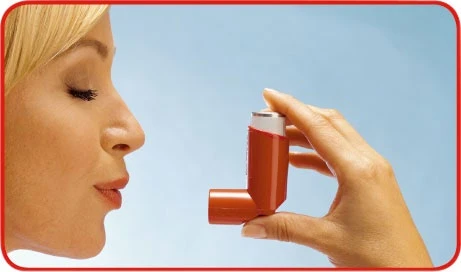 Perfil de una mujer rubia con los ojos cerrados, sosteniendo con la mano un inhalador naranja dirigido hacia su boca sobre un fondo azul claro