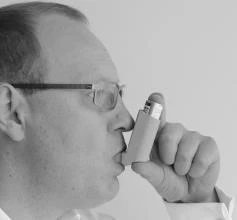 A man with glasses holds the inhaler to his mouth, preparing to take a dose of medication, shown in a black-and-white profile close-up