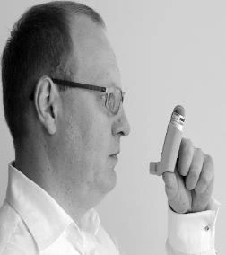 A man with glasses and wearing a white shirt holds an inhaler vertically in front of his face, ready to take a dose of medication