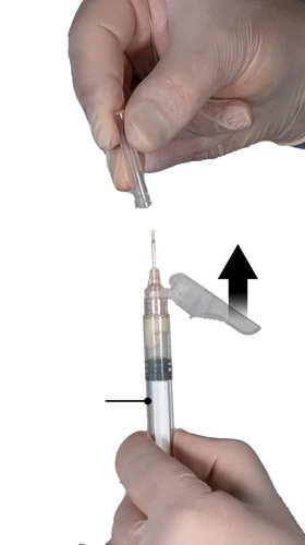 Mano con guante blanco retirando una protección transparente de la aguja de una jeringa llena de medicamento, indicado por una flecha negra y una línea de referencia