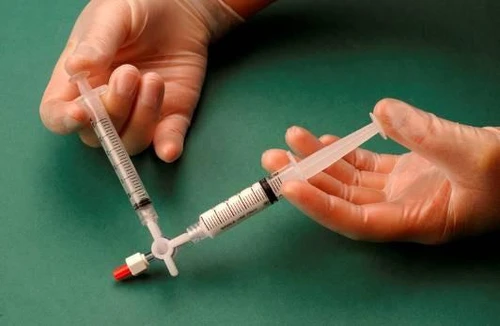 Due mani con guanti medici tengono delle siringhe collegate attraverso un rubinetto a tre vie bianco su sfondo verde
