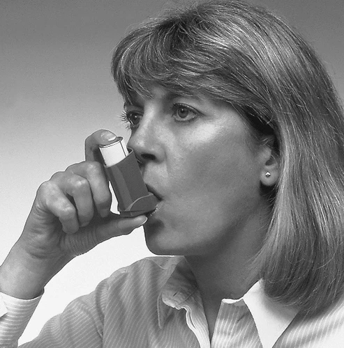 A woman in a light-colored shirt holds the inhaler to her mouth, preparing to take a dose of medication, shown in side profile against a light background