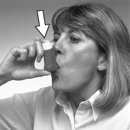 A woman holds the inhaler to her mouth, preparing to take the medication, with a white arrow pointing at the top part of the device