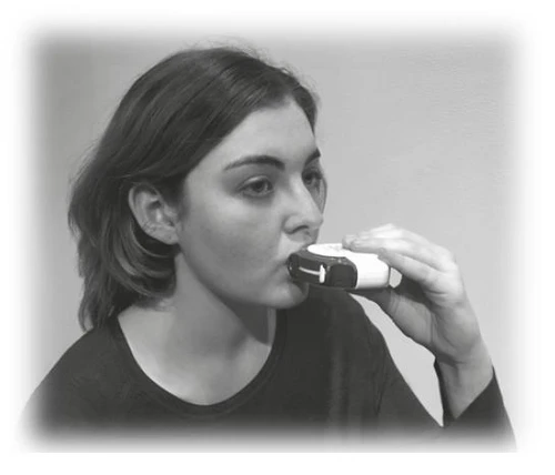 Kobieta trzyma biały inhalator przy ustach, przygotowując się do przyjęcia dawki leku, czarna koszulka, tło jednolite, czarno-biały kadr
