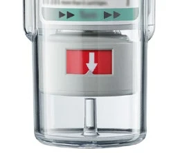 Close-up of a transparent medical dosing device with a white inner part and a red rectangle with a white downward-pointing arrow