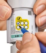 Hands holding a white auto-injector, with a blue pen marking one of the six boxes in a yellow control table with a blue checkmark